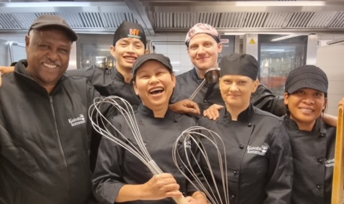 Sex glada personer med kockkläder och vispar i ett kök
